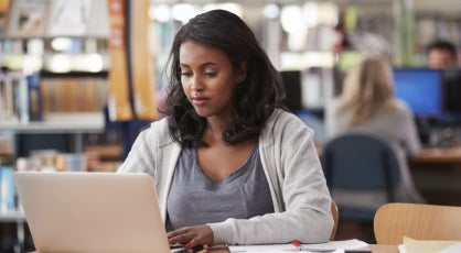 Woman working on a laptop in a public library, focused on her studies, suggesting career advancement or preparation for a finance manager role.