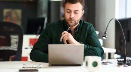 Man in a green shirt sits in a modern office at a desk with a laptop, holding a pen thoughtfully, suggesting focus on analytical work or career planning.