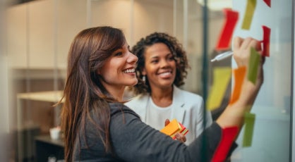 Two women smiling and collaborating while writing ideas on colorful sticky notes.