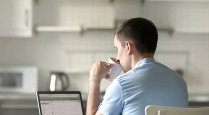Man in a light blue shirt sits at a kitchen table with a laptop, drinking from a cup, suggesting remote learning or online study in a home environment.