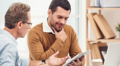 Two men in discussion at a desk, one holding a tablet and the other gesturing, with bookshelves and a laptop in the background, illustrating academic or educational consultation.