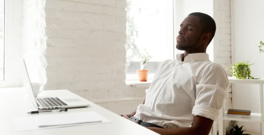 A man in a white shirt leans back at his desk in a bright, minimalist office with a laptop and potted plants, looking relaxed and contemplative.
