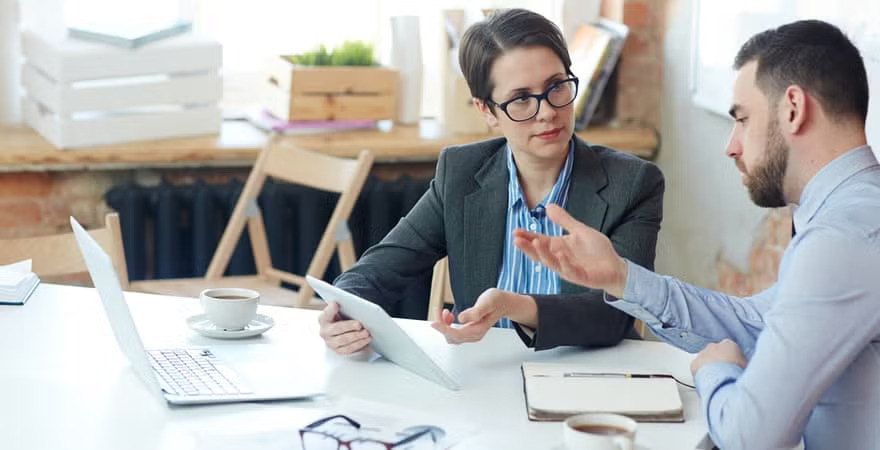 Two people in business attire are discussing something at a table with a laptop, tablet, notebook, and coffee cups in front of them.