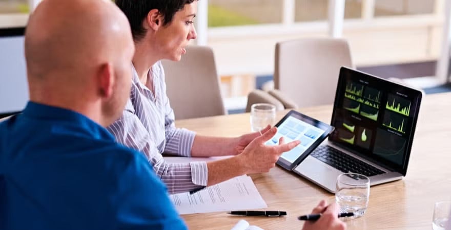 Two professionals sit at a table in a bright office, looking at digital analytics dashboards on a laptop and tablet while having a discussion.