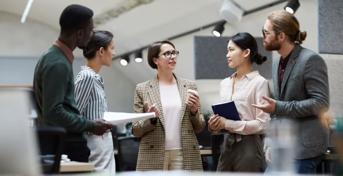 Group of five diverse professionals talking together in a modern office setting, suggesting workplace collaboration and communication.