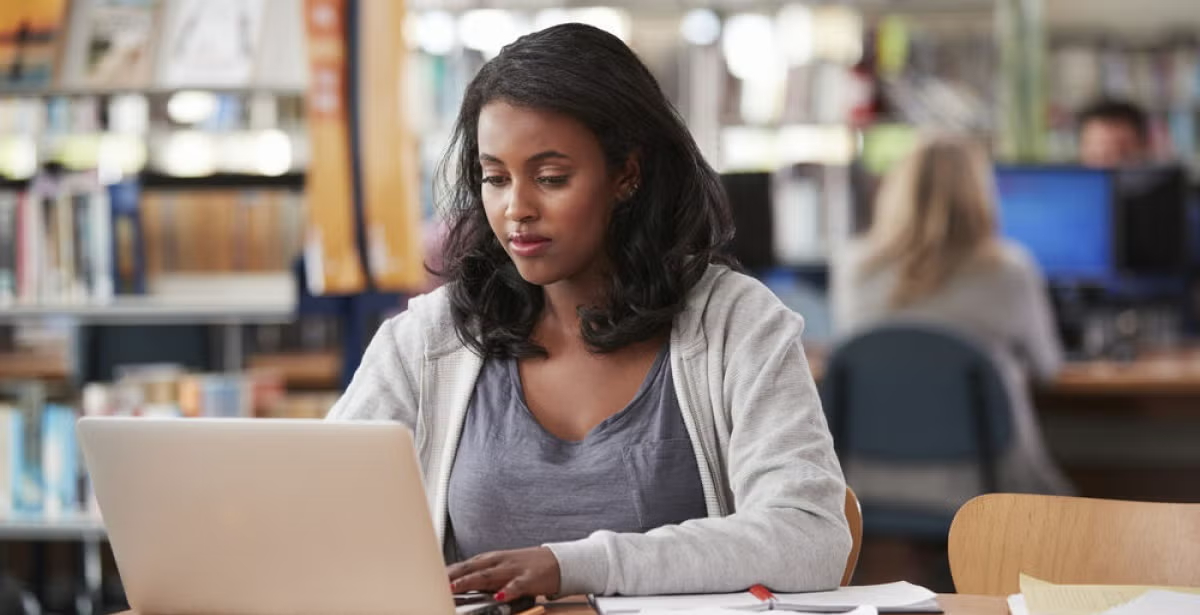 Woman working on a laptop in a public library, focused on her studies, suggesting career advancement or preparation for a finance manager role.