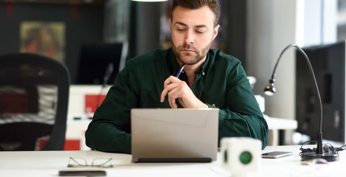 Man in a green shirt sits in a modern office at a desk with a laptop, holding a pen thoughtfully, suggesting focus on analytical work or career planning.