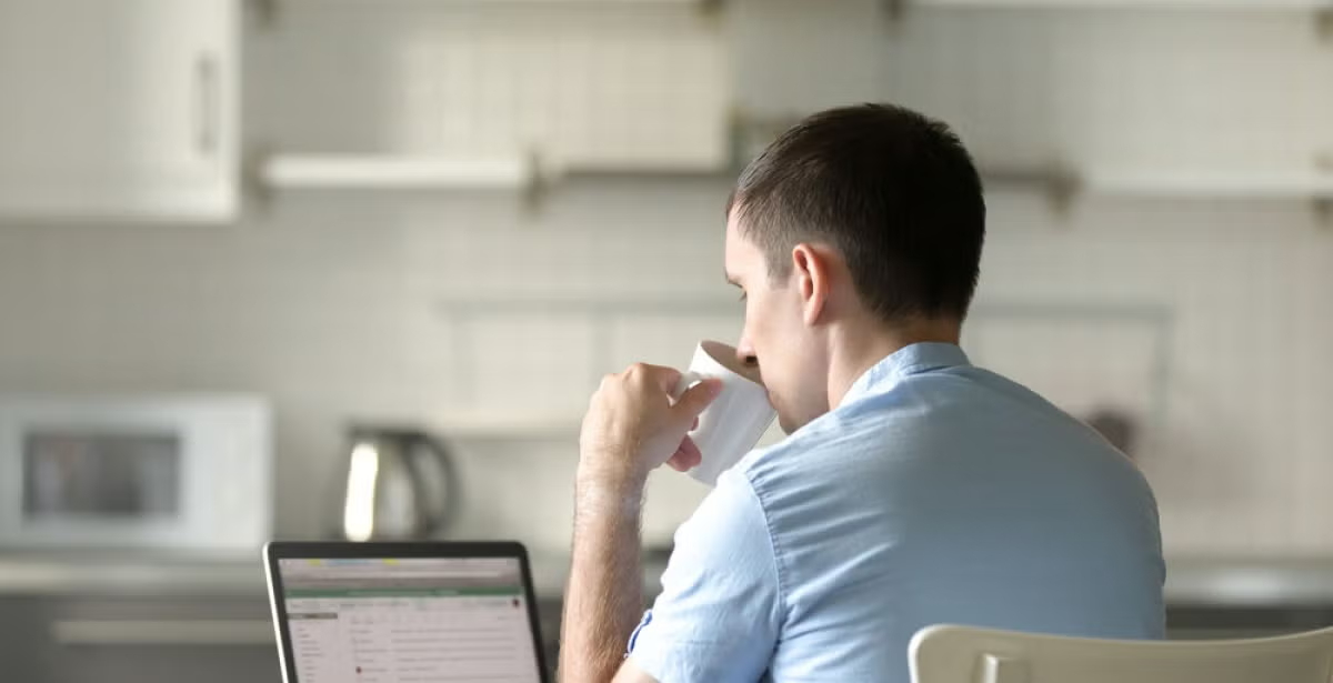Man in a light blue shirt sits at a kitchen table with a laptop, drinking from a cup, suggesting remote learning or online study in a home environment.