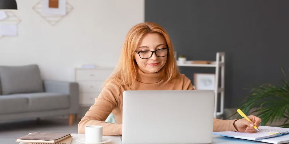 Woman in a tan sweater studies and takes notes with a laptop at a tidy desk, highlighting digital and applied learning.