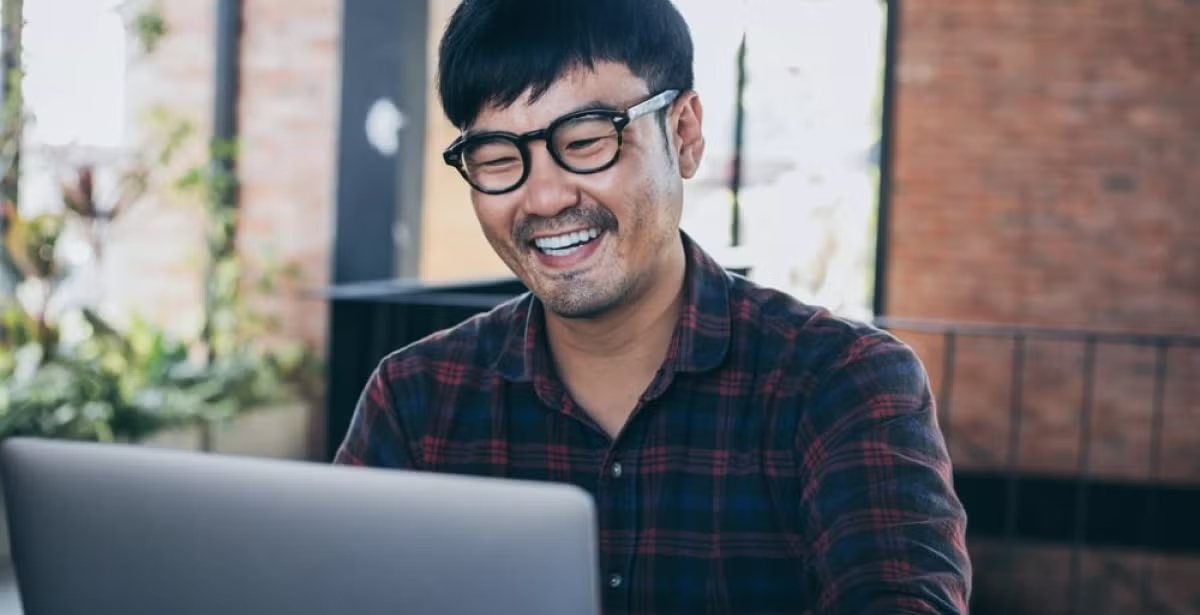 Man in glasses and a plaid shirt works on a laptop in a casual, modern work environment, conveying real-world financial application.