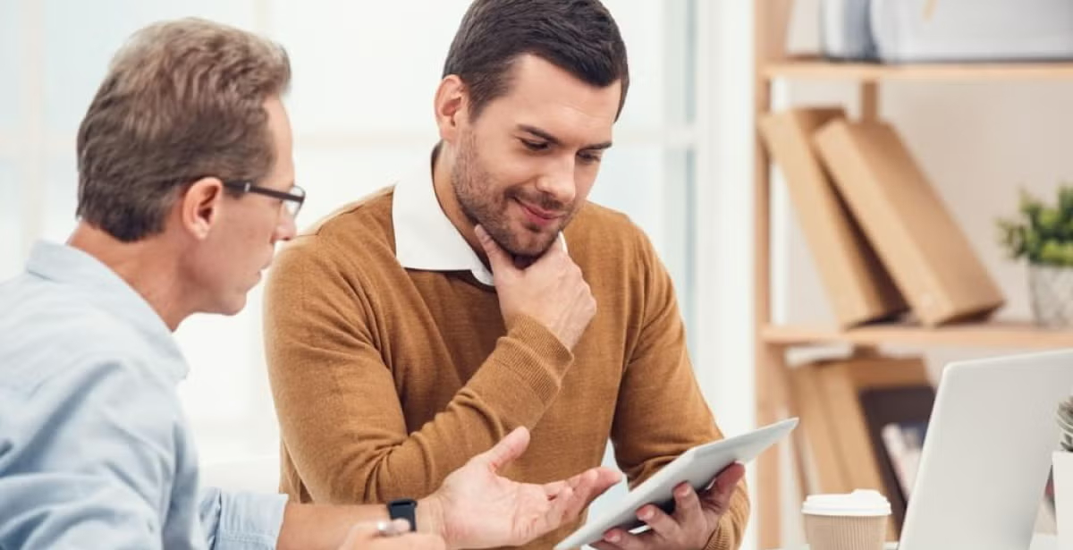 Two men in discussion at a desk, one holding a tablet and the other gesturing, with bookshelves and a laptop in the background, illustrating academic or educational consultation.
