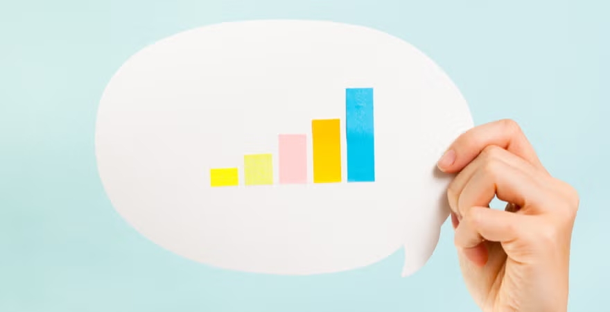 A person's hand holds a speech bubble cutout with multicolored bar charts, emphasizing the role of descriptive analytics in communicating trends and data findings.