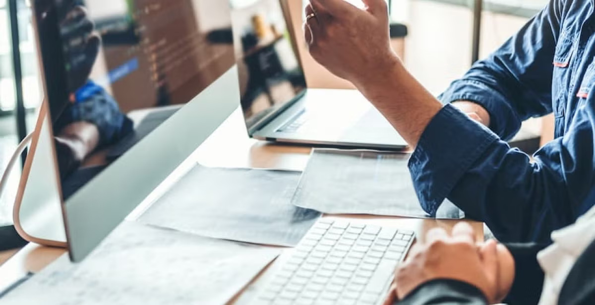 Two people working together at a desk with computers, papers, and keyboards, one person gesturing toward the screen while discussing something.